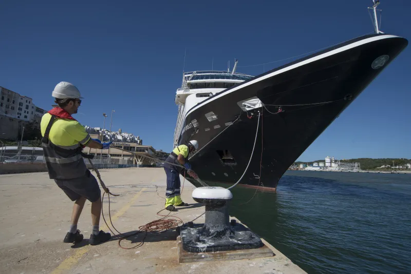 Dos trabajadores portuarios con casco y chaleco reflectante maniobran cuerdas de amarre para atracar un crucero negro de gran tamaño en el muelle del puerto, bajo cielo despejado