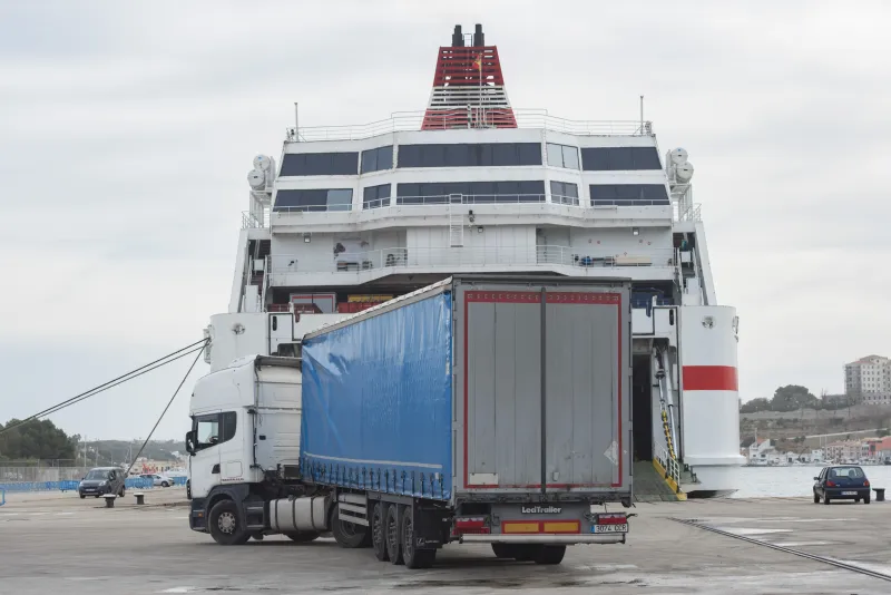 Camión de transporte con remolque azul se dispone a embarcar en un ferry mediante la rampa del muelle, con operarios portuarios y el casco urbano de Maó visible al fondo sobre la colina