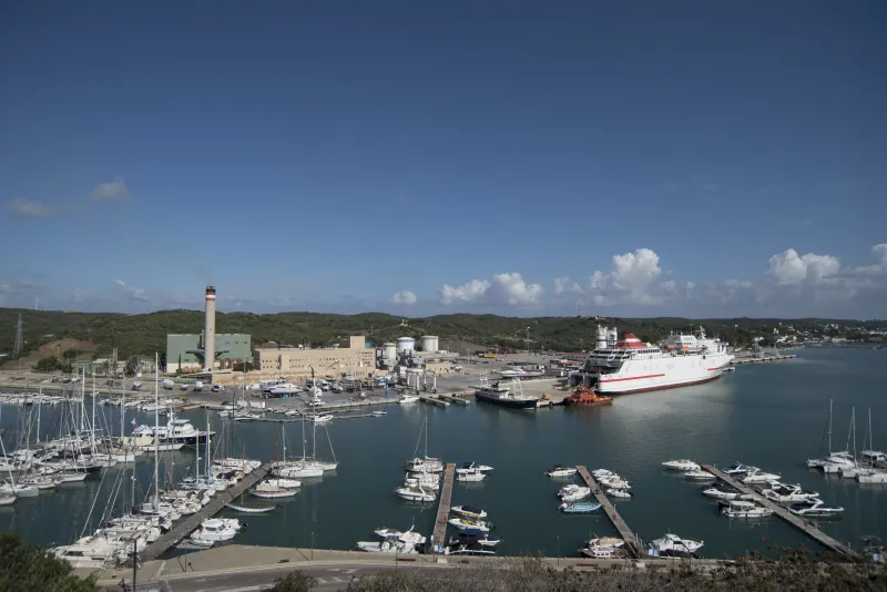 Panorámica del puerto de Maó con instalaciones industriales, una chimenea de central térmica y un ferry atracado junto a embarcaciones deportivas, bajo un cielo despejado