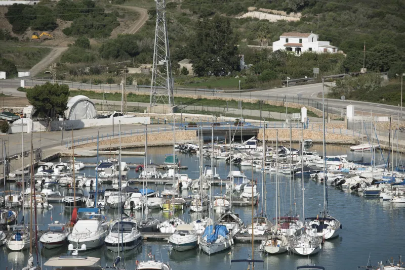 Embarcaciones de recreo atracadas en un muelle del puerto de Maó, junto a un pequeño puente peatonal que cruza un canal, rodeado de zonas verdes y carreteras