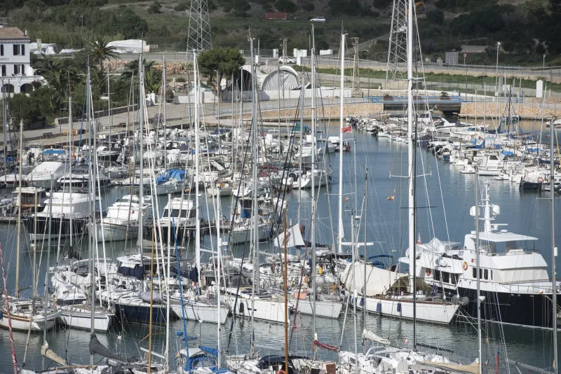 Numerosos veleros y yates atracados en el puerto de Maó, con mástiles que se alzan sobre el muelle y edificaciones portuarias visibles en segundo plano