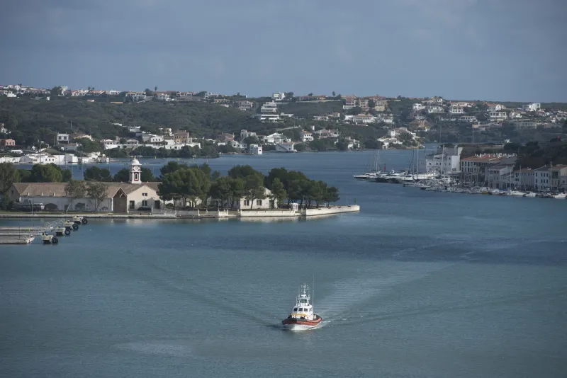 Una embarcación de práctico navega por el canal del puerto de Maó, rodeado de aguas tranquilas y edificaciones costeras. Al fondo se observan casas blancas en la colina y un pequeño conjunto de edificios históricos con torre y reloj junto a la orilla