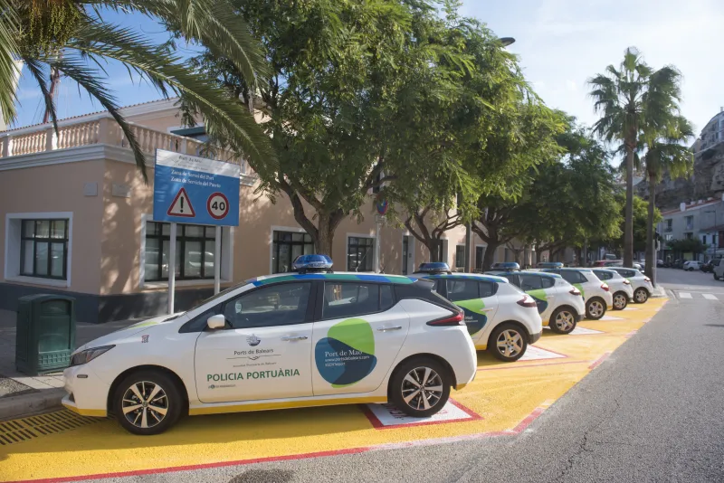 Varios vehículos eléctricos rotulados como “Policía Portuaria – Ports de Balears” estacionados en línea frente a un edificio administrativo del puerto, rodeado de árboles y señales de tráfico