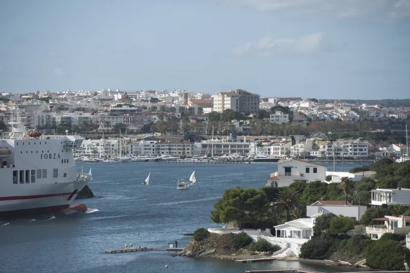 Panorámica del puerto de Maó con pequeñas embarcaciones a vela navegando frente a un buque de carga y el casco urbano de la ciudad extendido sobre la colina, con edificios históricos y modernos