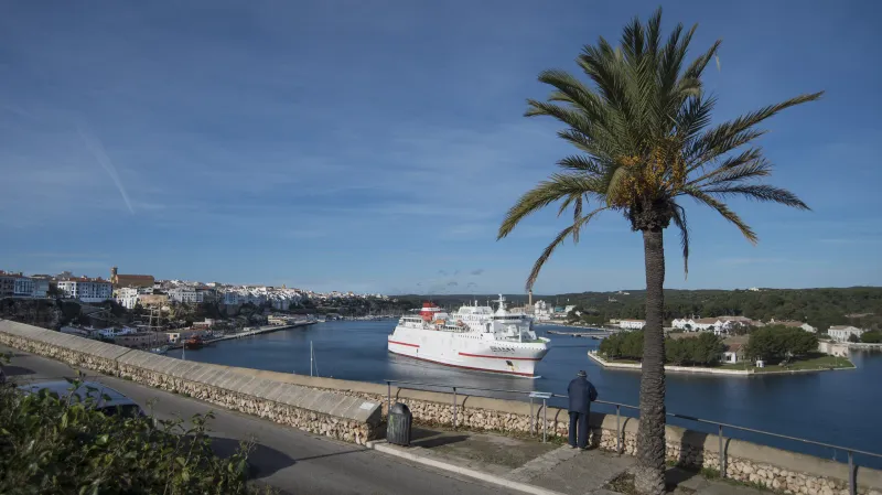 Un hombre se apoya en la barandilla del paseo marítimo de Maó observando un ferry blanco y rojo que navega por el canal. En primer plano, una palmera y una vía junto al acantilado; al fondo, el casco urbano y zonas verdes