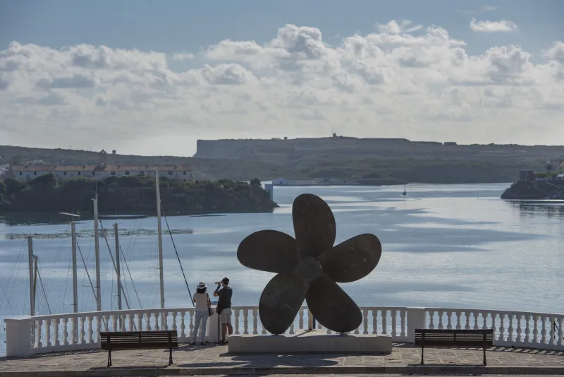 Vista panorámica del puerto de Maó desde un mirador con una gran hélice metálica expuesta como monumento; dos personas observan el paisaje marítimo y las antiguas fortificaciones al fondo