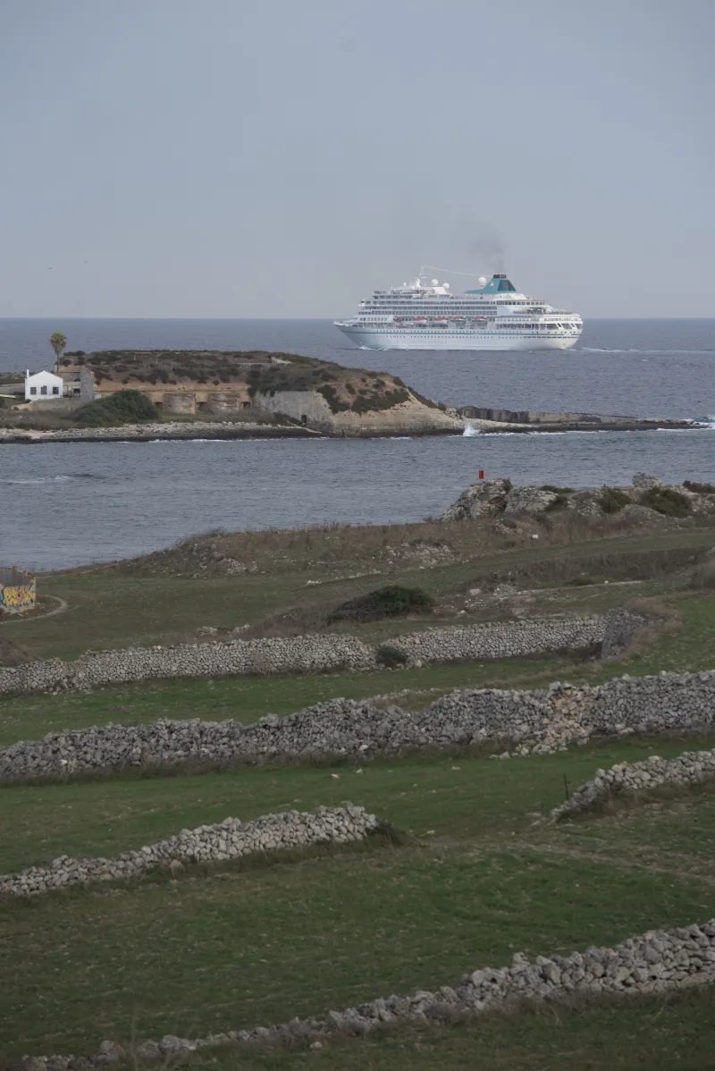 Paisaje del entorno costero del puerto de Maó con muros de piedra seca, vegetación baja y edificaciones antiguas junto al mar; al fondo se aprecia un buque de pasaje aproximándose a la bocana