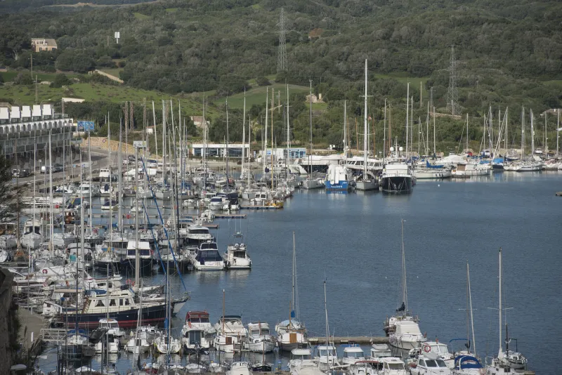 Vista general del puerto de Maó con numerosos barcos de recreo y veleros atracados en pantalanes, rodeados de colinas verdes y edificaciones portuarias al fondo.