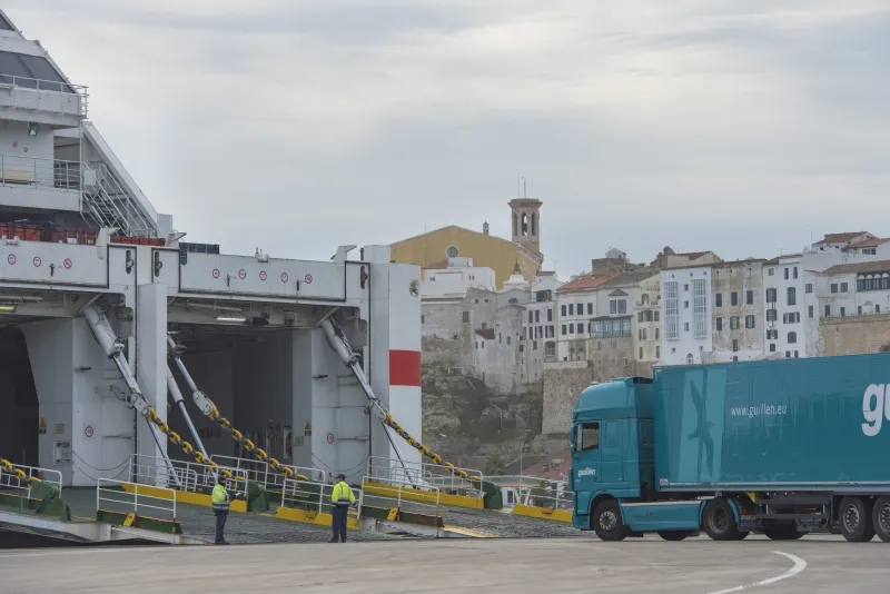 Camión articulado con remolque azul accediendo a la rampa trasera de un buque de pasajeros atracado en el puerto de Maó, en una operación de carga rodada