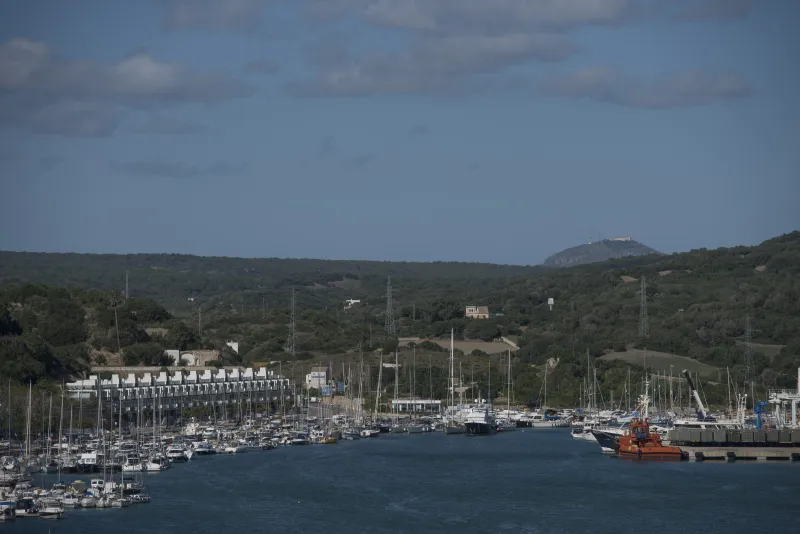 Imagen del puerto de Maó con un muelle ocupado por yates y barcos de recreo, rodeado de vegetación y colinas con edificaciones dispersas y una montaña visible en el horizonte