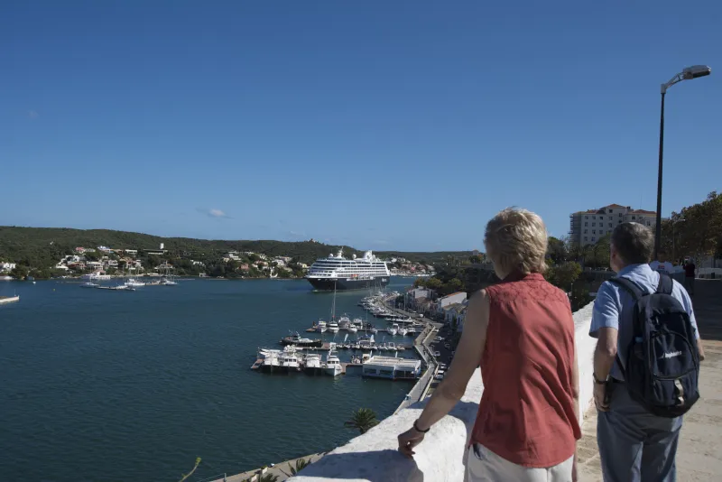 Dos personas observan desde un mirador el puerto de Maó, donde un gran crucero navega entre embarcaciones de recreo atracadas en el muelle. Al fondo se distinguen colinas verdes y edificaciones residenciales