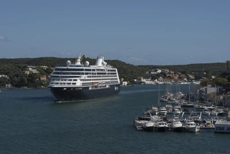 Un crucero de gran tamaño navega por el canal de entrada al puerto de Maó, flanqueado por pequeñas embarcaciones de recreo y viviendas en la costa, con colinas verdes al fondo