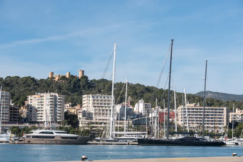 Vista del puerto de Palma con varios yates atracados frente al paseo marítimo; al fondo se alza el castillo de Bellver sobre una colina arbolada y tras él la ciudad con edificios residenciales modernos
