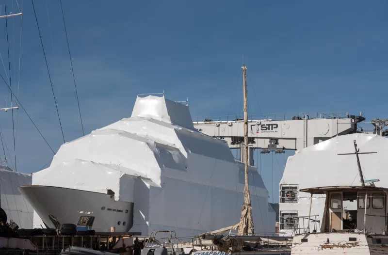 Grandes embarcaciones cubiertas con lonas protectoras blancas en una zona de mantenimiento del puerto de Palma; se observan grúas y estructuras metálicas de reparación junto a las instalaciones de STP Shipyard