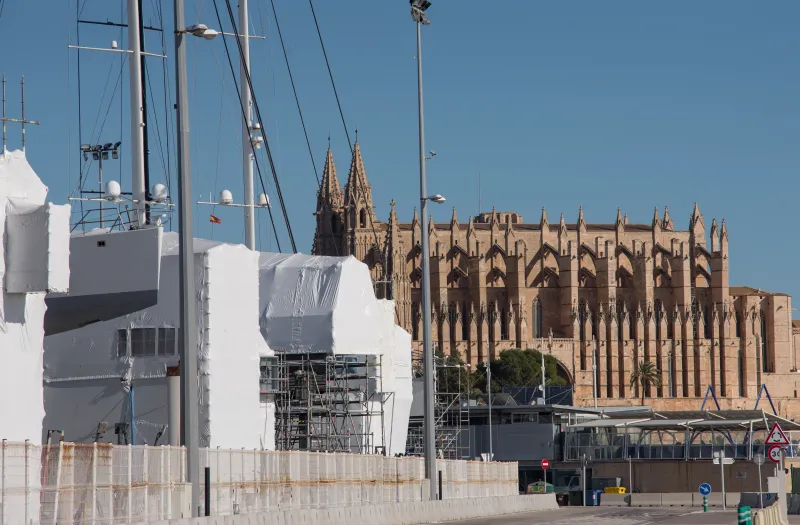 Embarcaciones de gran tamaño en proceso de mantenimiento en el puerto de Palma, con la Catedral de Santa María de Palma al fondo, destacando sus torres góticas sobre el casco urbano