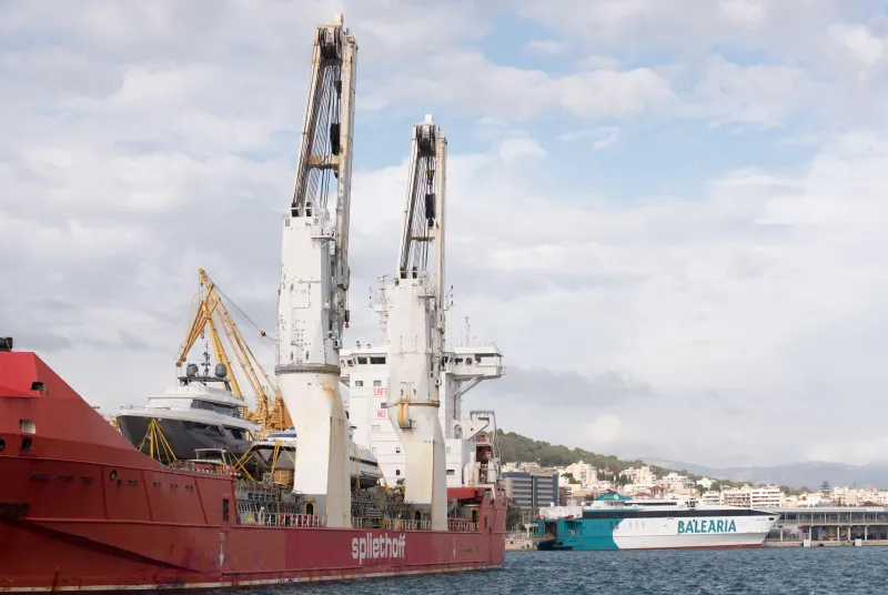 Grandes buques de carga y transporte atracados en el puerto, con grúas amarillas y blancas operando junto a las embarcaciones y un ferry de la compañía Baleària visible al fondo