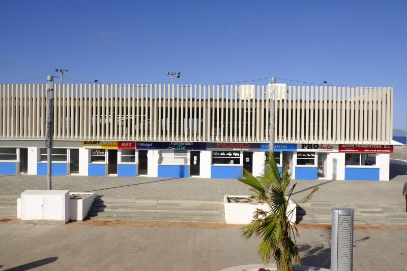 Fachada del edificio principal del puerto de La Savina con locales de alquiler de vehículos y servicios turísticos, entre ellos Avis, Budget y Formentera Mar, en una mañana soleada