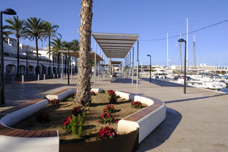 Paseo marítimo del puerto de La Savina con jardineras, bancos y pérgolas metálicas que proyectan sombra, bordeando la zona de atraque de embarcaciones deportivas