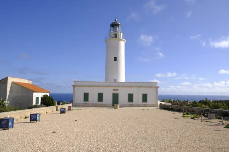 El faro de La Mola en Formentera se alza sobre una explanada abierta con el mar al fondo. El edificio blanco y el torreón central destacan bajo un cielo azul con nubes dispersas