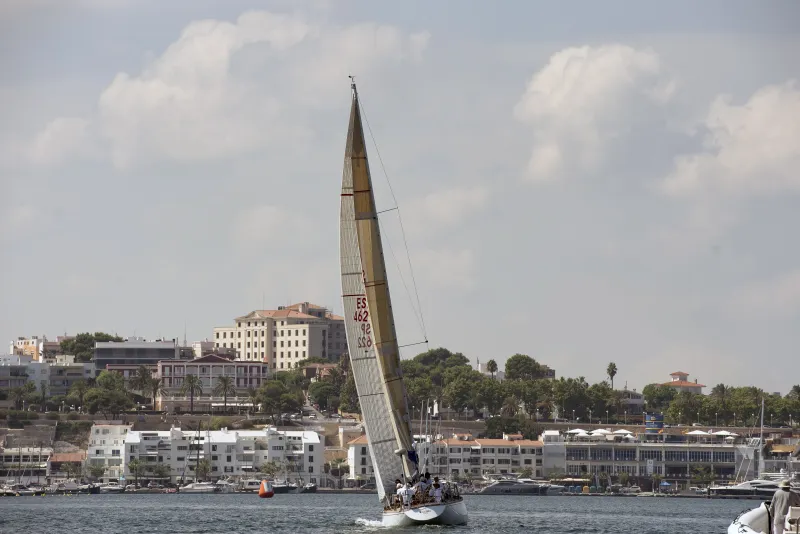 Velero de regata con tripulación navegando por las aguas del puerto de Maó, con el casco urbano al fondo y edificios históricos sobre la ladera que desciende hacia el mar